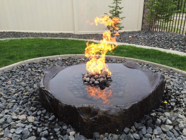 Water feature with fire added in the center laying flat in garden bed filled with basalt rock